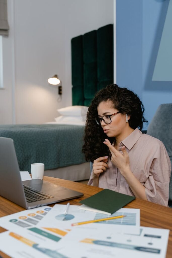 Woman working from home on a video call using a laptop, sitting at a desk in a modern bedroom.